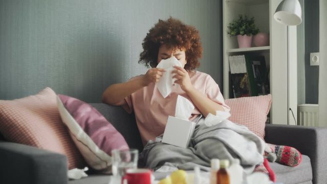 Sick african american girl with flu virus on sofa at home. She uses wipes to sneeze