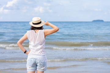 Fototapeta na wymiar Outdoor summer portrait of Young Asian woman wearing stylish hat and clothes standing on the beach, enjoying looking view of sea with blue sky on summer vacation.