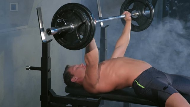 Professional muscular athletic man bodybuilder doing barbell chest exercise on bench in gym, side view. Man pumping chest with naked torso. Perfect bench press technique. White smoke in gym in dark.