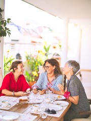 Meeting of middle age women having lunch and drinking coffee. Mature friends smiling happy using smartphone at home on a sunny day