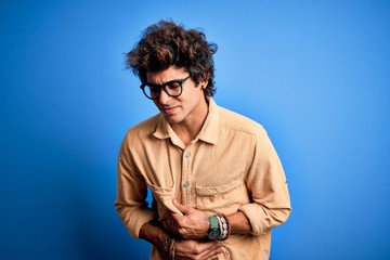 Young handsome man wearing casual shirt standing over isolated blue background with hand on stomach because nausea, painful disease feeling unwell. Ache concept.