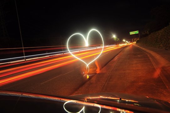 Blurred Motion Of Woman And Heart Shape Light Painting On Road Seen Through Car Windshield At Night