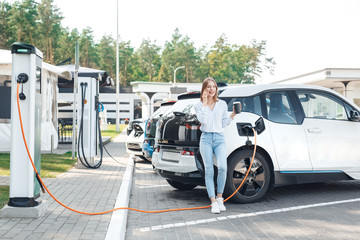 Transportation. Young woman on electric car having stop at charging station leaning on vehicle...
