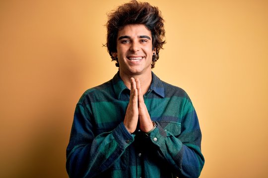 Young handsome man wearing casual shirt standing over isolated yellow background praying with hands together asking for forgiveness smiling confident.