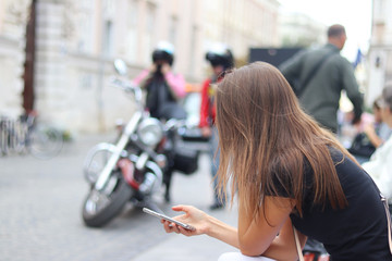 Naklejka na ściany i meble A young girl is looking into a smartphone that is holding. A woman sits on a bench in the center of an ancient city against a motorcycle and passers-by in a blurred blur. Lifestyle and leisure