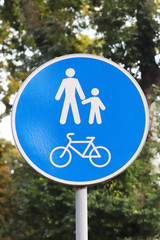 The round-shaped road sign is a pedestrian and bicycle zone against a background of green foliage. White people sign and bicycle on a blue background. Road and traffic safety. Symbols of urban areas