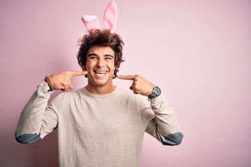 Young handsome man holding easter rabbit ears standing over isolated pink background smiling cheerful showing and pointing with fingers teeth and mouth. Dental health concept.