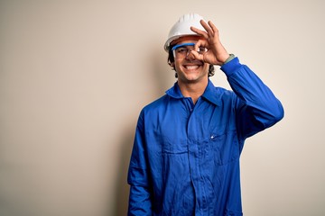 Young constructor man wearing uniform and security helmet over isolated white background doing ok gesture with hand smiling, eye looking through fingers with happy face.