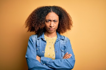 Young african american woman with afro hair wearing casual denim shirt over yellow background skeptic and nervous, disapproving expression on face with crossed arms. Negative person.