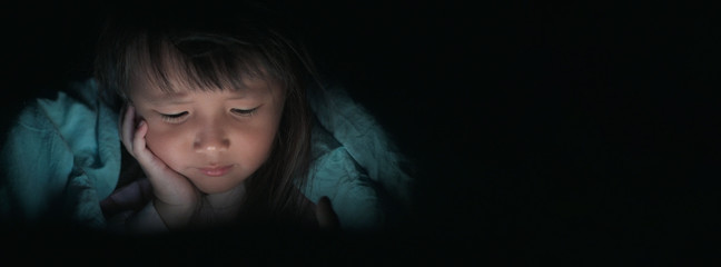 Little Girl watching tablets in bed at night with light flashes reflected from the screen,children using phones or playing tablet games and cartoon addiction smart phone concepts.wide banner.