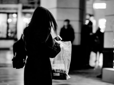 Blurred Unrecognizable Figure Of A Young Girl Hurrying Down Street In Dark Coat And A Mobile Phone In Her Hands. Stylized Silhouette Of A Young Modern Woman With A Backpack And A Package In Her Hands