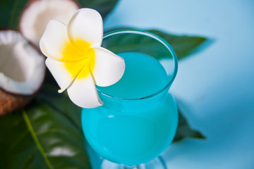 Exotic tropical Blue Curacao cocktail drink in a glass with Plumeria frangipani flower, palm leaf, fresh coconut on the background.