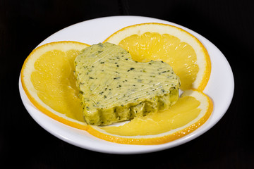 A piece of butter in the form of a heart with slices of orange on a white plate on a dark background