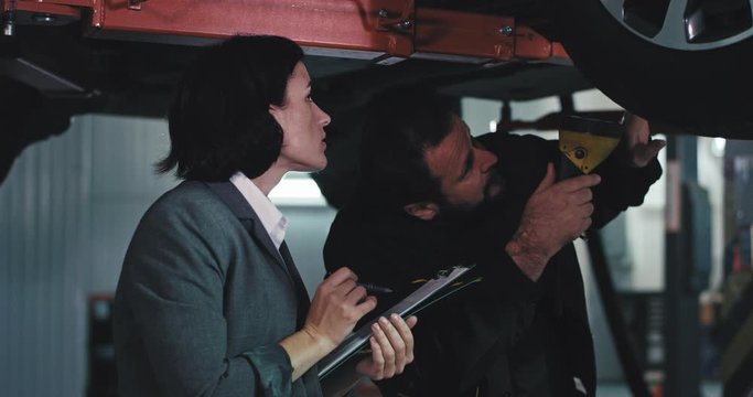 In a modern auto service center main mechanic mature man and the office manager woman in a suit they check the damaged car manager make some notes on her plan of work