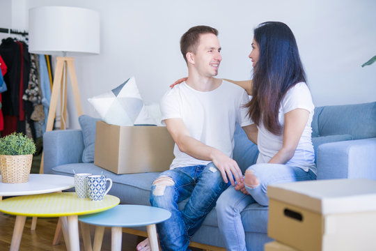 Young beautiful couple sitting on the sofa drinking coffee at new home around cardboard boxes