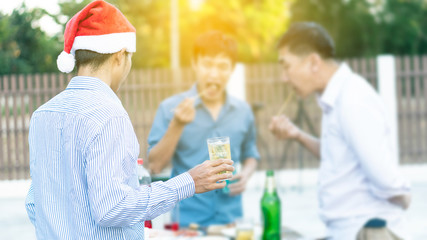 One man holds a glass of beer and two men are eating barbecue behind him and the orange light.
