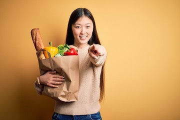 Young asian woman holding paper bag of fresh healthy groceries over yellow isolated background pointing to you and the camera with fingers, smiling positive and cheerful