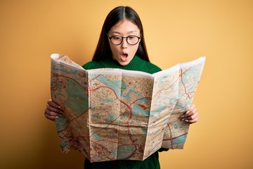 Young asian turist woman looking at city tourist map on a trip over yellow background scared in shock with a surprise face, afraid and excited with fear expression