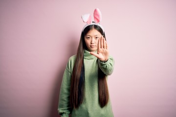 Young asian woman wearing cute easter bunny ears over pink background doing stop sing with palm of the hand. Warning expression with negative and serious gesture on the face.