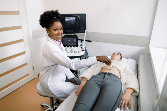 Young black woman doctor sonographer using ultrasound machine at work, holding ultrasound scanner in hand while examine female patient abdomen at hospital.