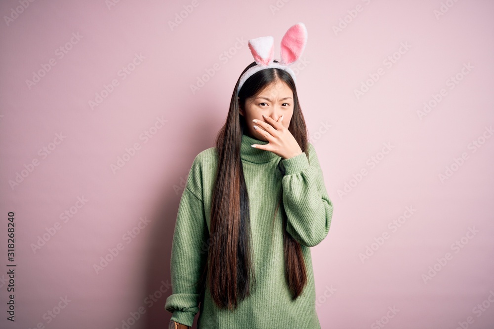 Poster Young asian woman wearing cute easter bunny ears over pink background smelling something stinky and disgusting, intolerable smell, holding breath with fingers on nose. Bad smell