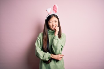 Young asian woman wearing cute easter bunny ears over pink background looking confident at the camera with smile with crossed arms and hand raised on chin. Thinking positive.