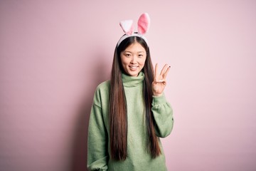 Young asian woman wearing cute easter bunny ears over pink background showing and pointing up with fingers number three while smiling confident and happy.