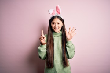 Young asian woman wearing cute easter bunny ears over pink background showing and pointing up with fingers number six while smiling confident and happy.