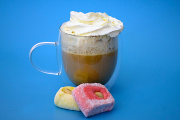 Cappuccino with whipped cream on a blue background. Double bottom cup with coffee and pink rahat lakoum candies. Tasty hot drink in a transparent cup.