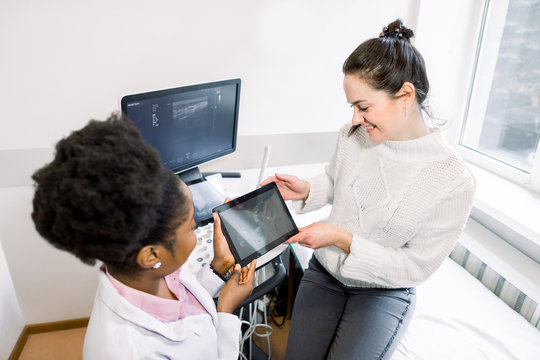 Young Caucasian pregnant woman and African woman doctor looking together at baby ultrasound picture, holding digital tablet