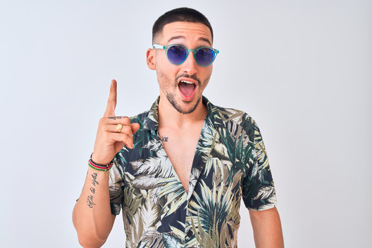 Young handsome man wearing Hawaiian sumer shirt and blue sunglasses over isolated background pointing finger up with successful idea. Exited and happy. Number one.