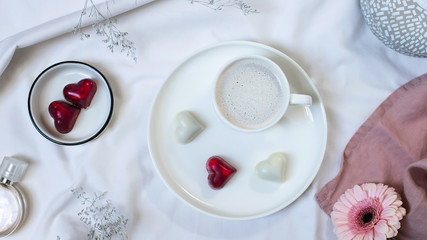 cup of coffee with cream, bonbons and flowers on a white bed. breakfast in bed for your beloved wife. flat lay, top view, banner