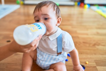 Adorable toddler sitting on the floor drinking milk using feeding bottle at kindergarten