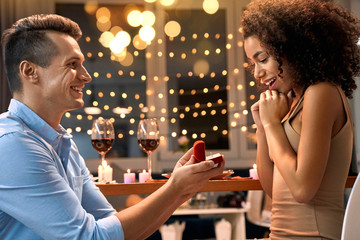 Romantic Date. Young multiethnic couple at home having dinner man standing on knee proposing to woman with proposal ring smiling surprised