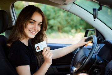 Smiling young female with pleasant appearance shows proudly her drivers license, sits in new car,...