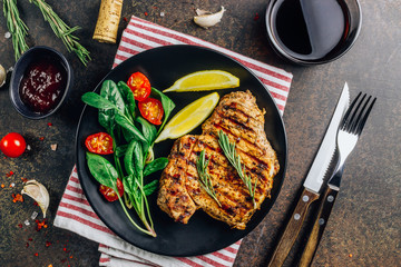 Roasted pork steak on black plate over dark background. With lemon, Cranberry Sauce and wine.