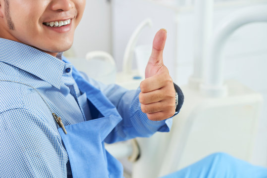 Unrecognizable Asian Man Wearing Protective Bib Sitting On Dental Chair Smiling And Showing Thumb Up, Horizontal Shot