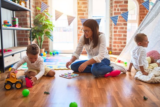 Beautiful teacher and toddler playing with tractor around lots of toys at kindergarten