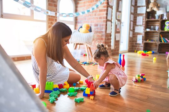 Young beautiful teacher and toddler sitting on the floor playing with building blocks toy at kindergarten