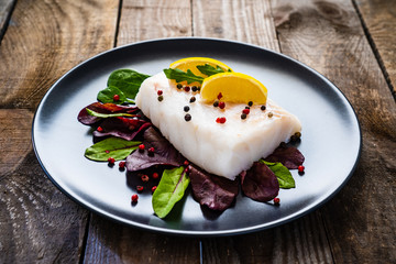 Fresh raw cod with vegetable leaves and lemon  on black plateon on wooden table