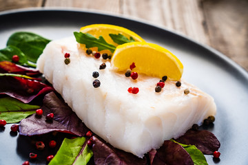 Fresh raw cod with vegetable leaves and lemon  on black plateon on wooden table