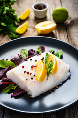 Fresh raw cod with vegetable leaves and lemon  on black plateon on wooden table