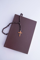 A wooden cross with a rosary on a closed Holy Bible. Closed book with a brown cover and a symbol of christians on white background.