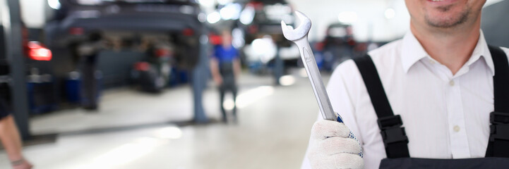 Obraz premium Portrait of cheerful man holding shining metal wrench and standing in modern car maintenance garage with many automobiles. Machinery repairman and service station concept