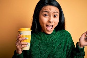 Young beautiful chinese woman drinking cup of coffee over isolated yellow background screaming proud and celebrating victory and success very excited, cheering emotion