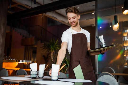 Young happy waiter lays the table in cafe