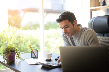 A young man working at a desk with a laptop. Calculator, notepad, pencil, pen and cup of coffee on the side.