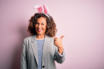 Middle age beautiful woman wearing bunny ears standing over isolated pink background smiling with happy face looking and pointing to the side with thumb up.