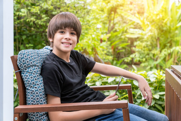 Young cute Caucasian boy using digital tablet sitting on couch in garden at home, looking at camera