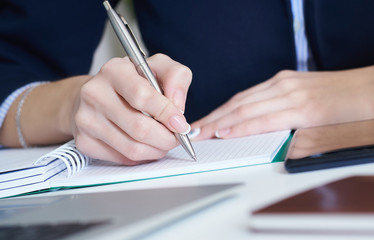 Young businesswoman writing in notepad while sitting at the office. Female hands holding a pen and making notes close up.